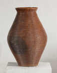 Brown ribbed pottery vase with tall narrow neck and burnished finish on white pedestal against plain background.