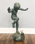 Bronze child statue balancing on one leg above large bronze snail, garden fountain against light-colored wall.