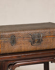 Brown rattan and iron Qing Dynasty suitcase trunk on dark wooden table, plain backdrop, close-up.