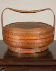 Gold and tan woven rattan Chinese basket with painted cover and arched handle on a table.