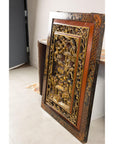 Gold giltwood Qing wall panel with carved figural court scene, leaning against a wall in a bright room.