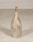 Cream and brown drip-glazed ceramic bottle vase on a plain light-colored floor, vertical view.