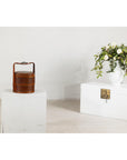 Brown vintage woven carrying box with double handles on white pedestal, next to a chest and vase of flowers.