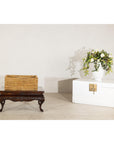 Brown rattan rectangular storage box with lid on wooden table beside a white trunk with flowers, country style setting.