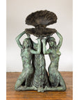 Bronze Greco Roman style three nymph fountain statue holding shell, displayed on a wooden floor.