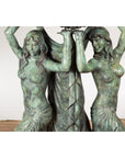Bronze Greco Roman style nymph fountain with three figures in flowing dresses, arms raised, set against a white brick wall.