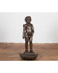 Dark patina bronze winged cherub statue on shell, close-up, displayed by a white brick wall and wood floor.