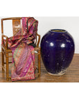 Pink and gold patterned fabric over wooden chair next to oversized cobalt blue glazed Thai ceramic vessel, interior setting.