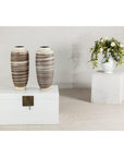 Close-up of two tall ceramic vases with horizontal swirl design on a white chest, flowers in white vase in background.
