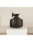 Close-up of a vintage hand-hammered metal jug with ring handle on a white pedestal against a plain background.