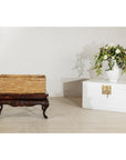 Close-up of a vintage rectangular handwoven rattan basket with side handles beside a white chest and floral arrangement.