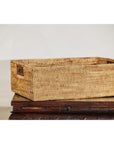 Brown handwoven rattan rectangular basket with side handles on carved wooden surface against a plain backdrop.
