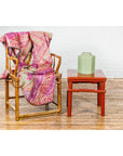 Pink patterned blanket on bamboo chair next to red square side table with humpback stretcher, styled interior setting.