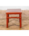 Red wooden square side table with humpback stretcher on a worn wooden floor, white brick wall background.