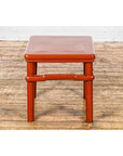 Red wooden square side table with humpback stretcher in front of a white brick wall.