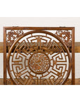 Brown wood fretwork panel with phoenix, bats, and geometric maze, displayed on woven table against white brick wall.