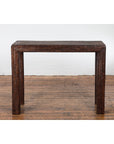 Dark brown woven rattan console table with four legs on wood floor, against a white brick wall.