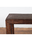 Close-up of a dark brown woven rattan console table on a wooden floor against a white wall.
