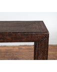 Close-up of dark brown woven rattan console table on wooden floor against a white brick wall.