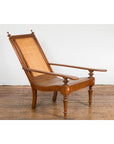 Brown wood and rattan Dutch Colonial lounge chair with carved finials, angled backrest, flat armrests on wooden floor.