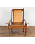 Brown wood and rattan Dutch Colonial lounge chair with carved finials on wooden floor against a white wall.
