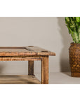 Distressed wood country style two-tier coffee table beside leafy plant in woven pot, plain wall background.