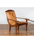 Brown leather Dutch Colonial plantation lounge chair with wide arms on wooden floor by a white brick wall.