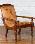 Brown leather Dutch Colonial plantation lounge chair on wooden floor against white brick wall, early 20th-century style.