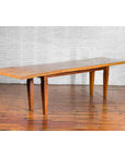 Brown mango wood Indonesian dining table with tapered legs on wooden floor, white brick wall background.