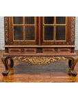 Gilded wood Dutch Colonial cabinet with bonnet top and glass doors on wooden floor against a brick wall.