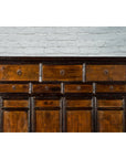 Brown wood Qing Dynasty armoire cabinet with 14 drawers, set against a light gray brick wall background.