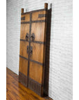Brown wooden Qing Dynasty palace doors with iron fittings, set against a white brick wall and wood floor.