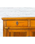 Close-up of brown elmwood Chinese side chest with bronze medallion hardware, set against a white brick wall.
