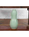 Green glazed ceramic gourd-shaped vase on wooden table against brick wall, close-up.