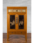 Black lacquered wooden armoire with gilt warrior motifs and two drawers, against a white brick background.
