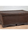 Dark brown rattan antique basket coffee table with lid on wooden floor against a white brick wall.
