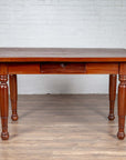 Brown teak antique Dutch Colonial Javanese desk with single drawer and turned legs, against white brick wall and wood floor.