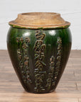 Green glazed Chinese pottery water jug with calligraphy, placed on a wooden floor next to a white wall.
