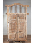 Natural wood bonnet top window balcony with hand-carved foliage motifs, set against a brick wall and wood floor.