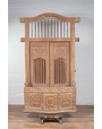 Natural wood bonnet top window balcony with hand-carved foliage motifs, white brick wall and wood floor background.