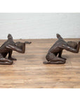 Vintage bronze kneeling table bases with raised arms on a wooden floor against a white wall.