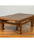 Brown distressed wood Indonesian coffee table with block legs on hardwood floor against a white brick wall.