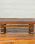 Brown distressed wood Indonesian coffee table with block legs on polished wood floor against a white wall.