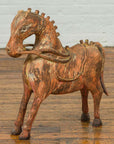 Rust-colored carved wooden horse figurine from Madras on a wooden floor with a white brick wall background.