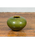 Olive green porcelain urn with circular design, glazed finish, displayed on wooden floor against white wall.