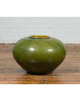 Olive green porcelain circular urn with glossy glaze on wooden floor, positioned by a white brick wall.