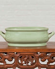 Green celadon ceramic foot bath with two handles and foliage motifs on ornate wooden table, light background.