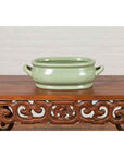 Green celadon ceramic vintage foot bath with two handles and foliage motifs on wooden table in front of brick wall.