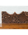 Brown carved teak floral panel on wooden cabinet, set against a white brick wall.