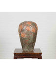 Close-up of orange and green ceramic Thai garden vase with vine leaf motifs on wooden stand against white brick wall.